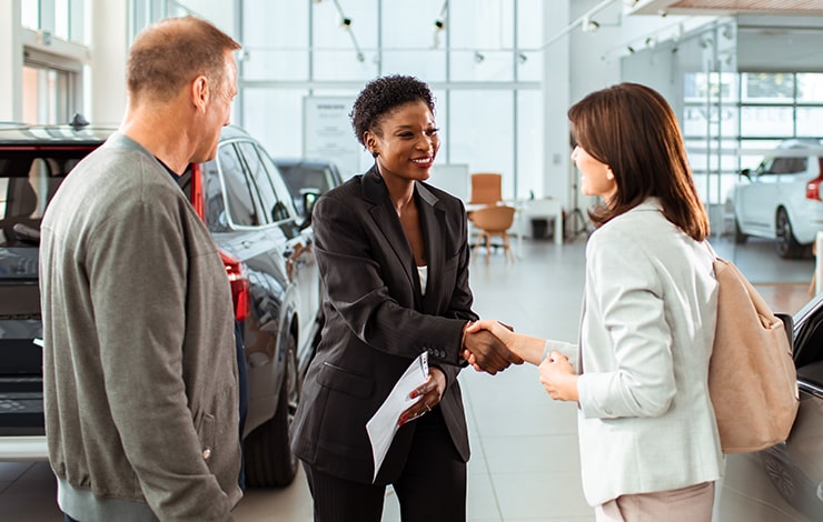 Car saleswoman shaking the hand of a woman while a man stands next to her in a car dealership