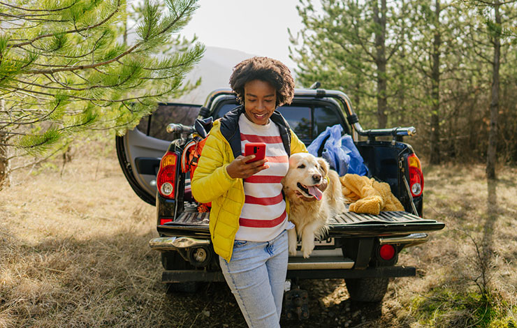 A woman is smiling and looking at her phone while hugging a dog. The woman and dog are sitting on the back of a truck in the woods.