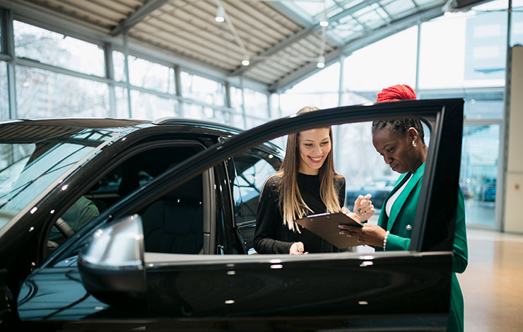 Saleswoman and a female customer in a car dealership. Sales manager looking at clipboard and explaining the car features to woman customer in showroom.