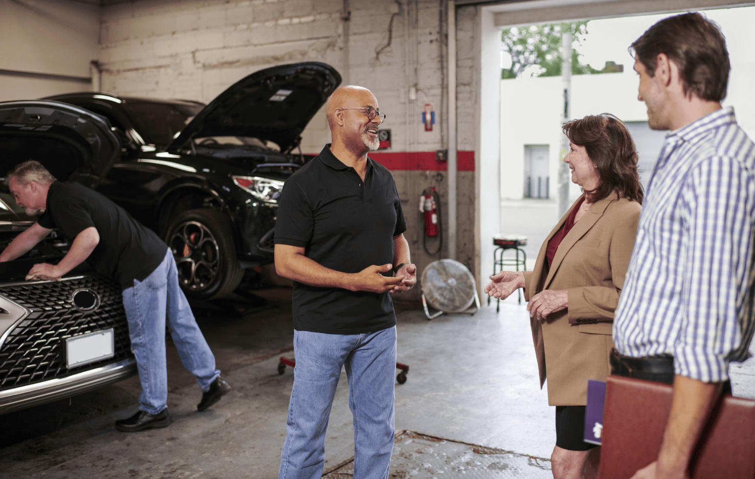 A group of people are smiling and talking in an auto body garage. A man is standing in the background actively working on the engine of a car.