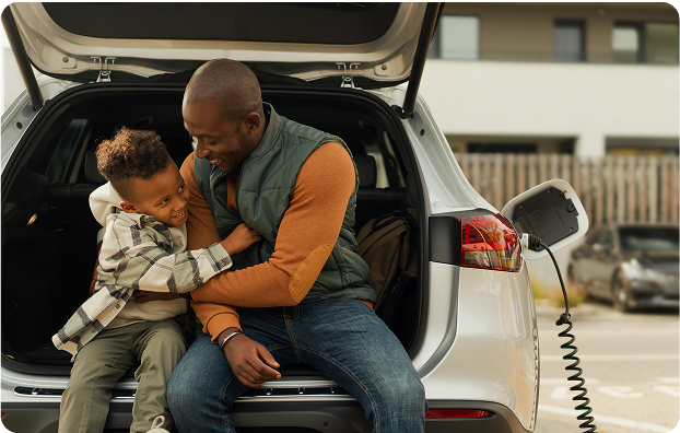 A father and a young son sit in the back of their electric vehicle playing while the car charges